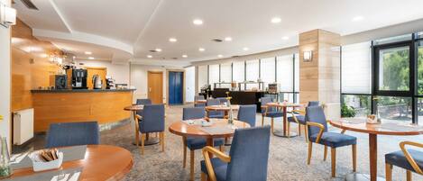 A modern hotel lobby with round tables and blue chairs, a reception desk, and large windows offering a view of the outdoors.
