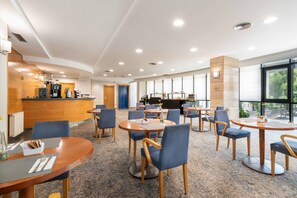 A modern hotel lobby with round tables and blue chairs, a reception desk, and large windows offering a view of the outdoors. - Exe Area Central (Santiago de Compostela)