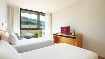 Een hotelkamer met twee bedden, een bureau met een laptop en een raam met uitzicht op groen.