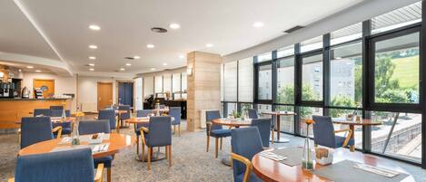 A modern hotel lobby with round tables, blue chairs, a reception desk, and large windows offering a view of the outdoors.
