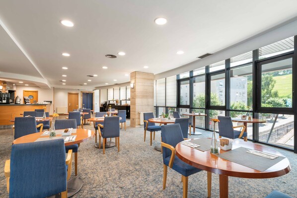 Un moderno vestíbulo de hotel con mesas redondas, sillas azules, una recepción y amplios ventanales con vistas al exterior.