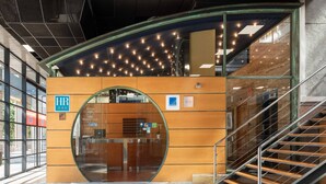 A modern reception area with a glass entrance, a wooden reception desk, and a staircase leading to an upper level. - Exe Area Central (Santiago de Compostela)