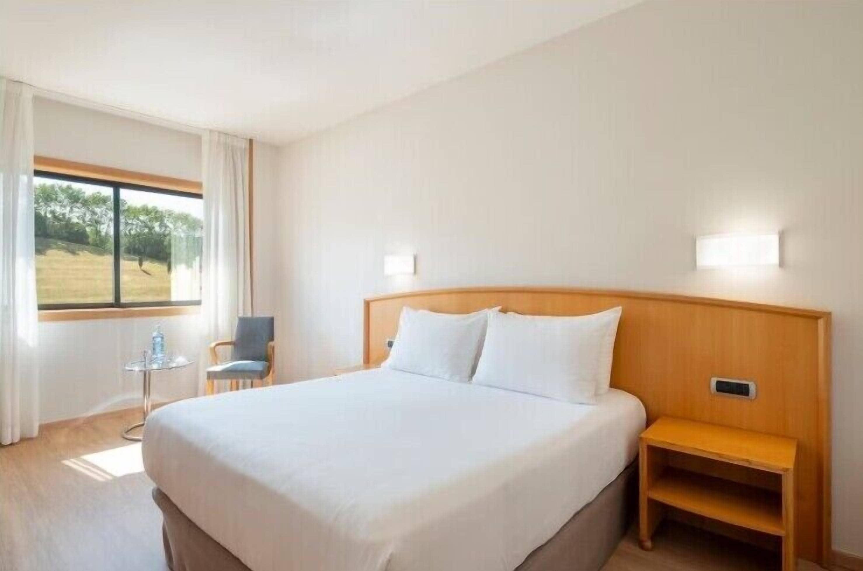Habitación de hotel con una cama grande, una mesita de noche, una silla, una pequeña mesa y una ventana con cortinas.