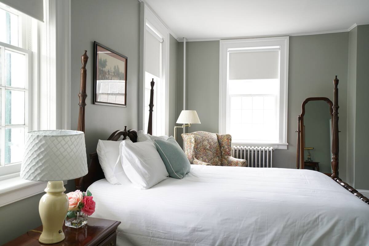 Un dormitorio ordenado con una cama grande, una mesita de noche con lámpara, una silla y un cuadro en la pared.
