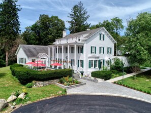 A large white building with green shutters, a covered patio with red umbrellas, and a well-maintained garden. - Le Chambord at Curry Estate (Hopewell Junction)