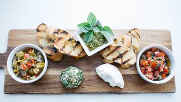 A wooden board with grilled bread, a bowl of vegetable salad, a bowl of tomato salsa, a ball of herb cheese, and a small bowl of pesto.