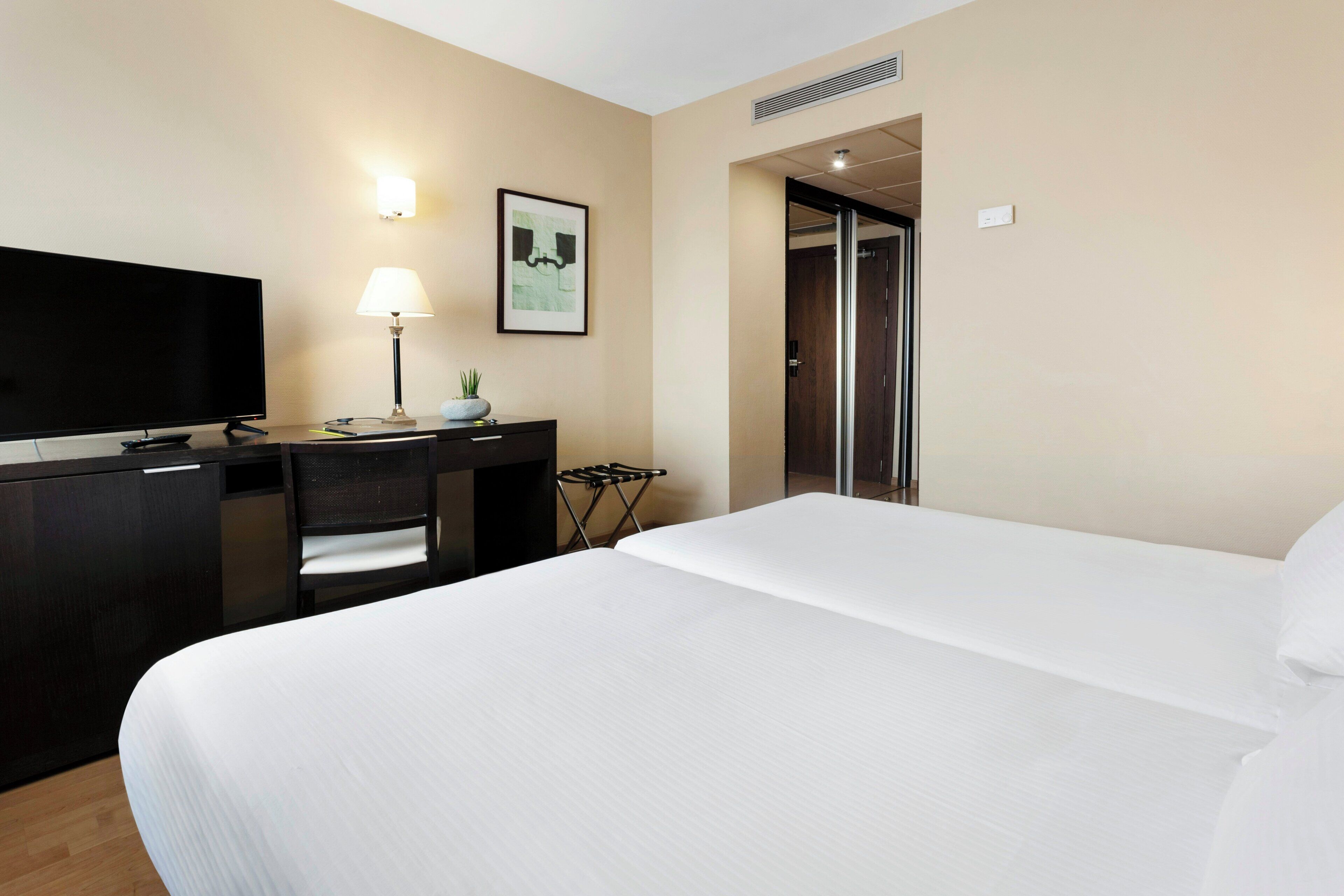 A hotel room with a bed, a desk with a television, a lamp, a framed picture, and a door.