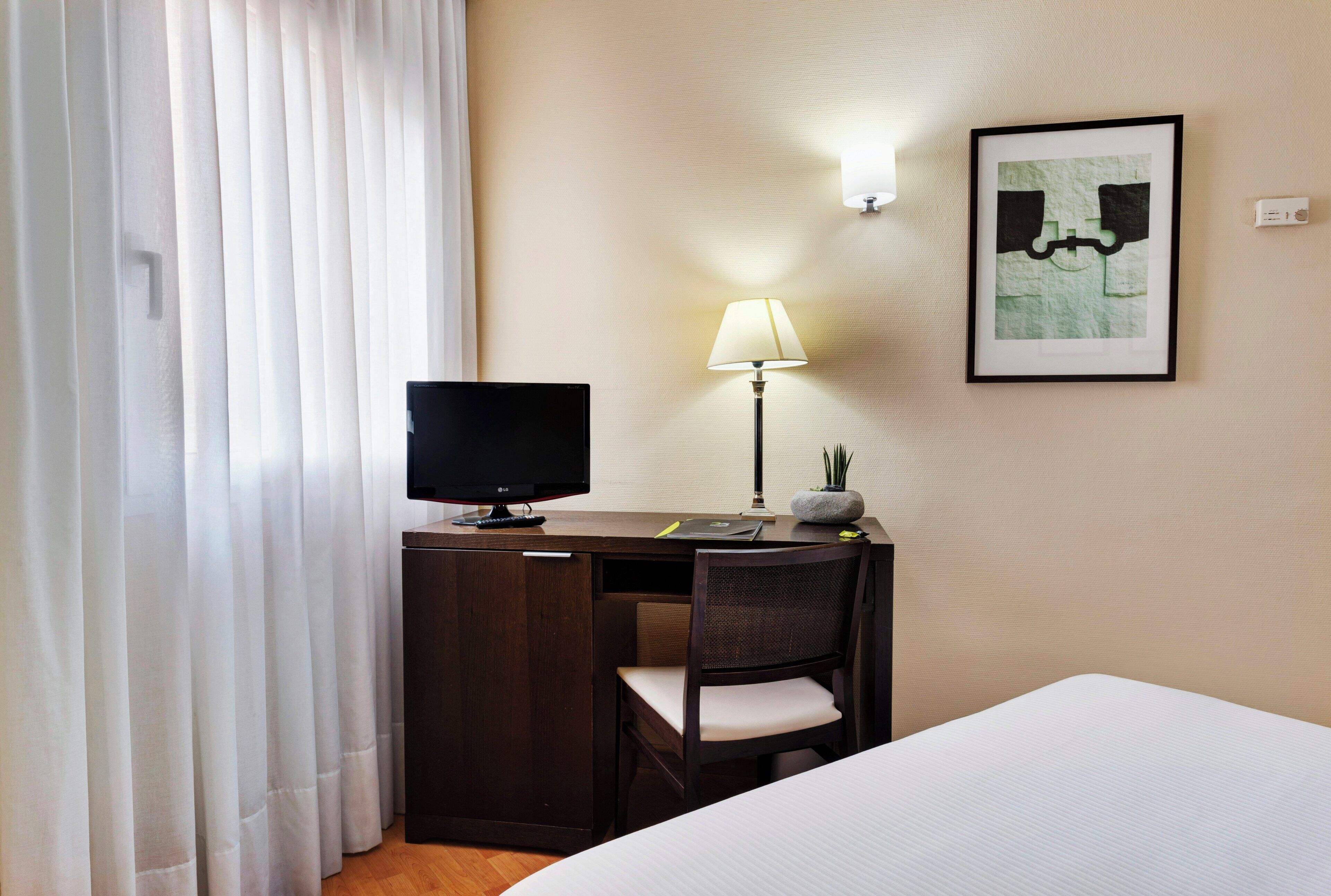 A hotel room with a bed, a desk with a computer monitor, a chair, a lamp, and a framed picture on the wall.