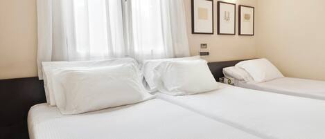 A hotel room with two beds, white linens, a window with sheer curtains, and framed artwork on the wall.