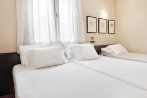 Una habitación de hotel con dos camas, ropa de cama blanca, una ventana con cortinas traslúcidas y cuadros en la pared.
