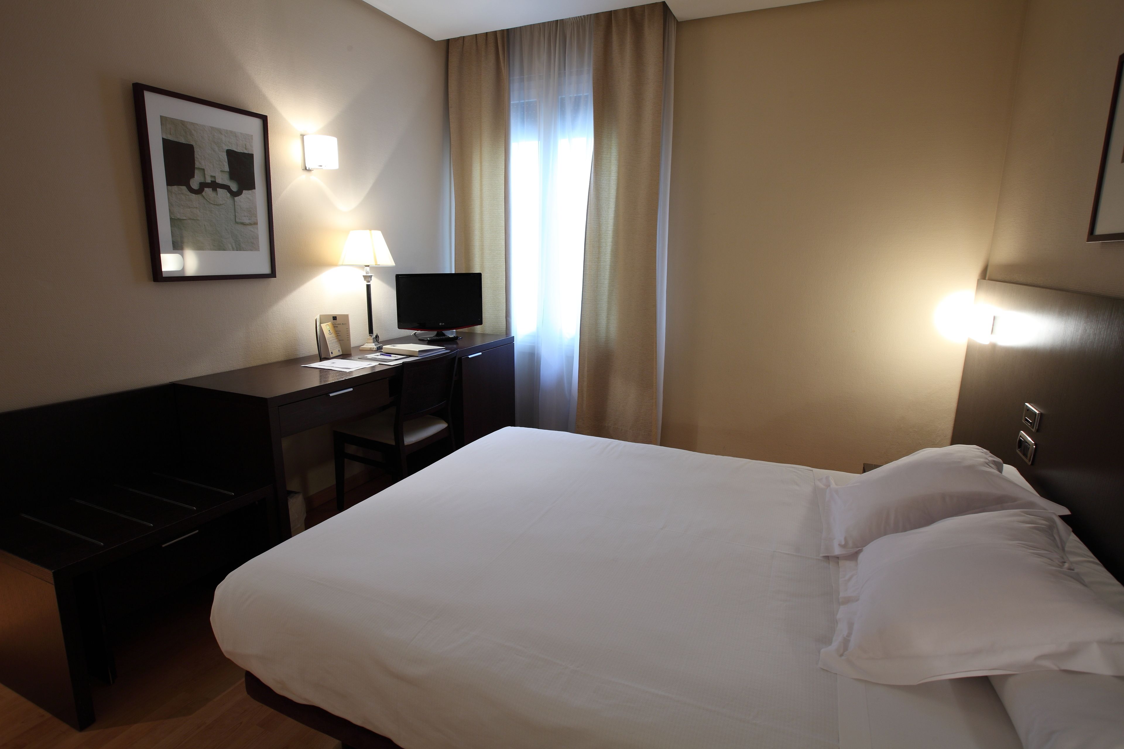 Una habitación de hotel con cama, escritorio, silla, televisor y un cuadro en la pared.