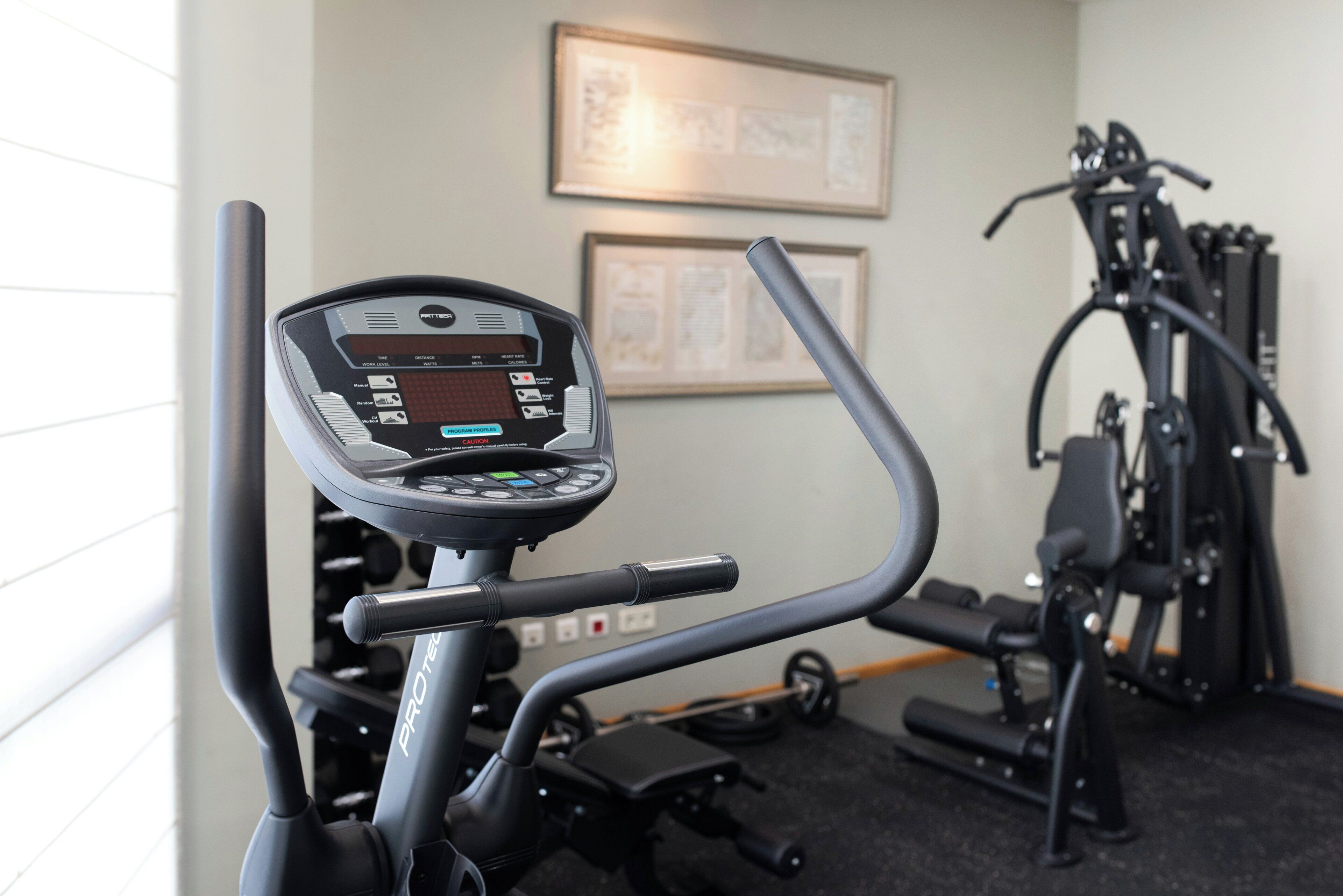 Equipamento de ginástica com visor digital e suporte de halteres fixado na parede.