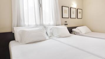 Een hotelkamer met twee bedden, witte lakens, een raam met dunne gordijnen en twee ingelijste foto's aan de muur.