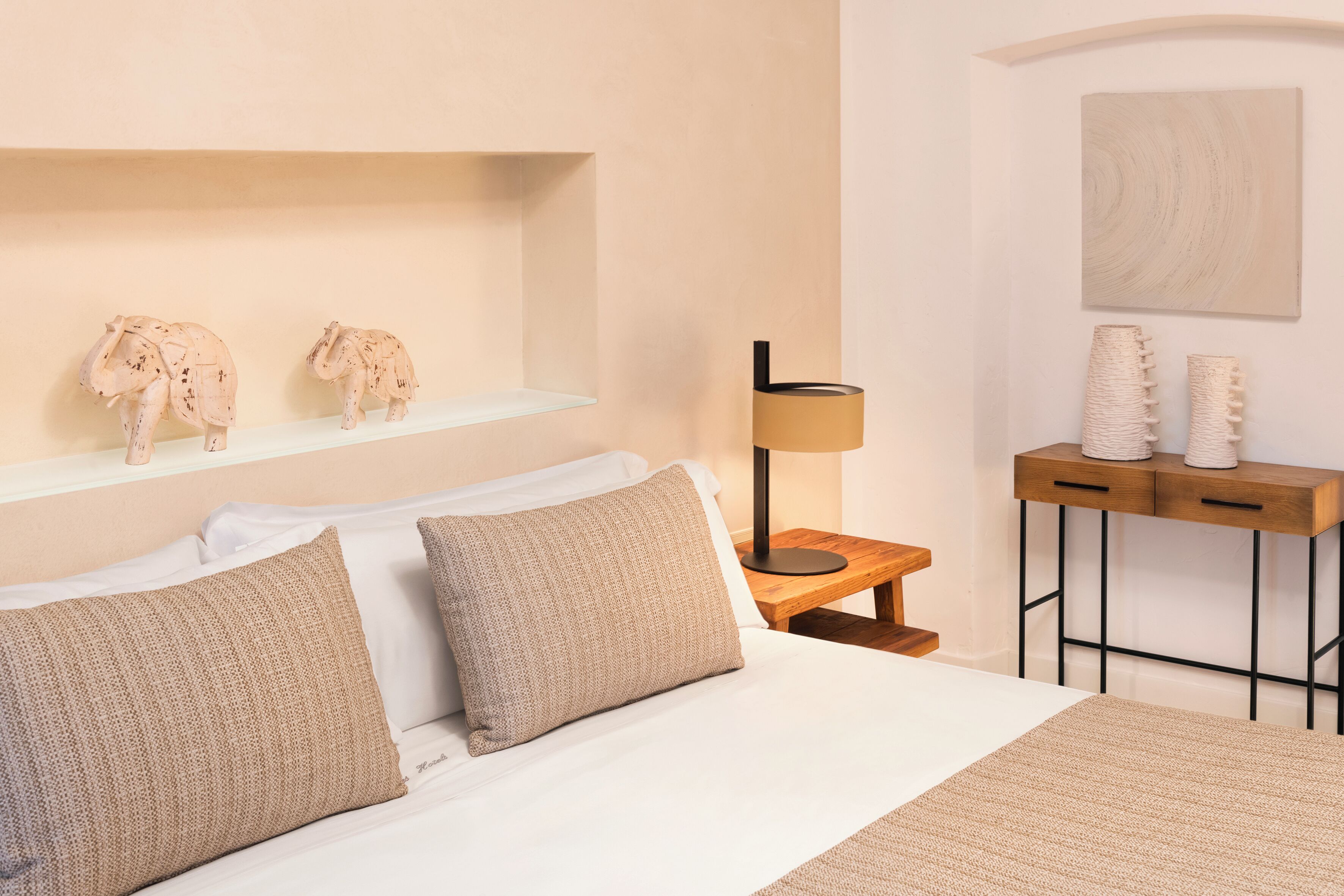 Un dormitorio ordenado con cama, almohadas, una mesita de noche, lámpara y elementos decorativos en la pared.