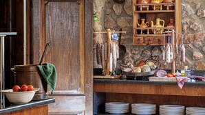 Une cuisine rustique avec un mur en pierre, des étagères en bois ornées de poteries, un porte-viande suspendu et une porte.