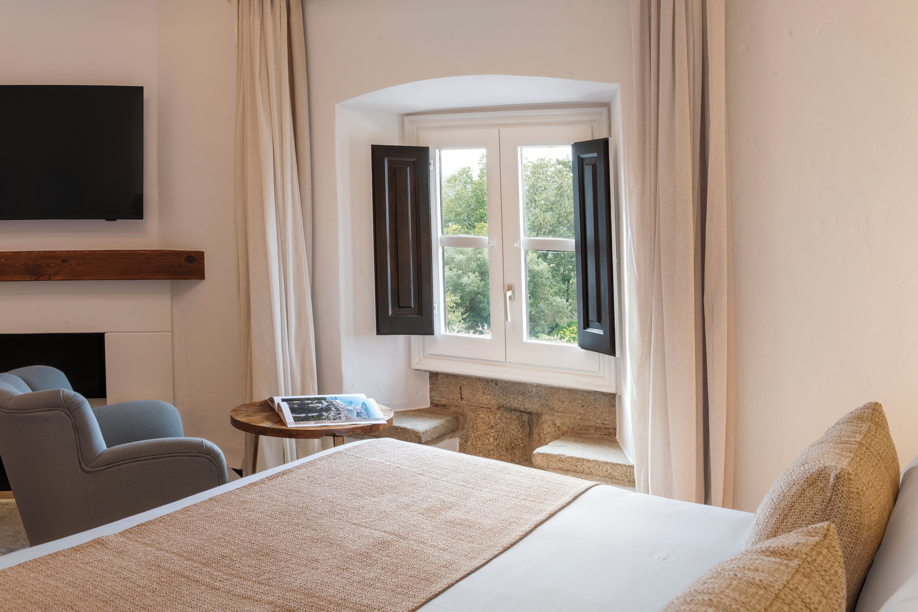 A hotel room with a bed, a TV mounted on the wall, a small table with a book, and a window with curtains.