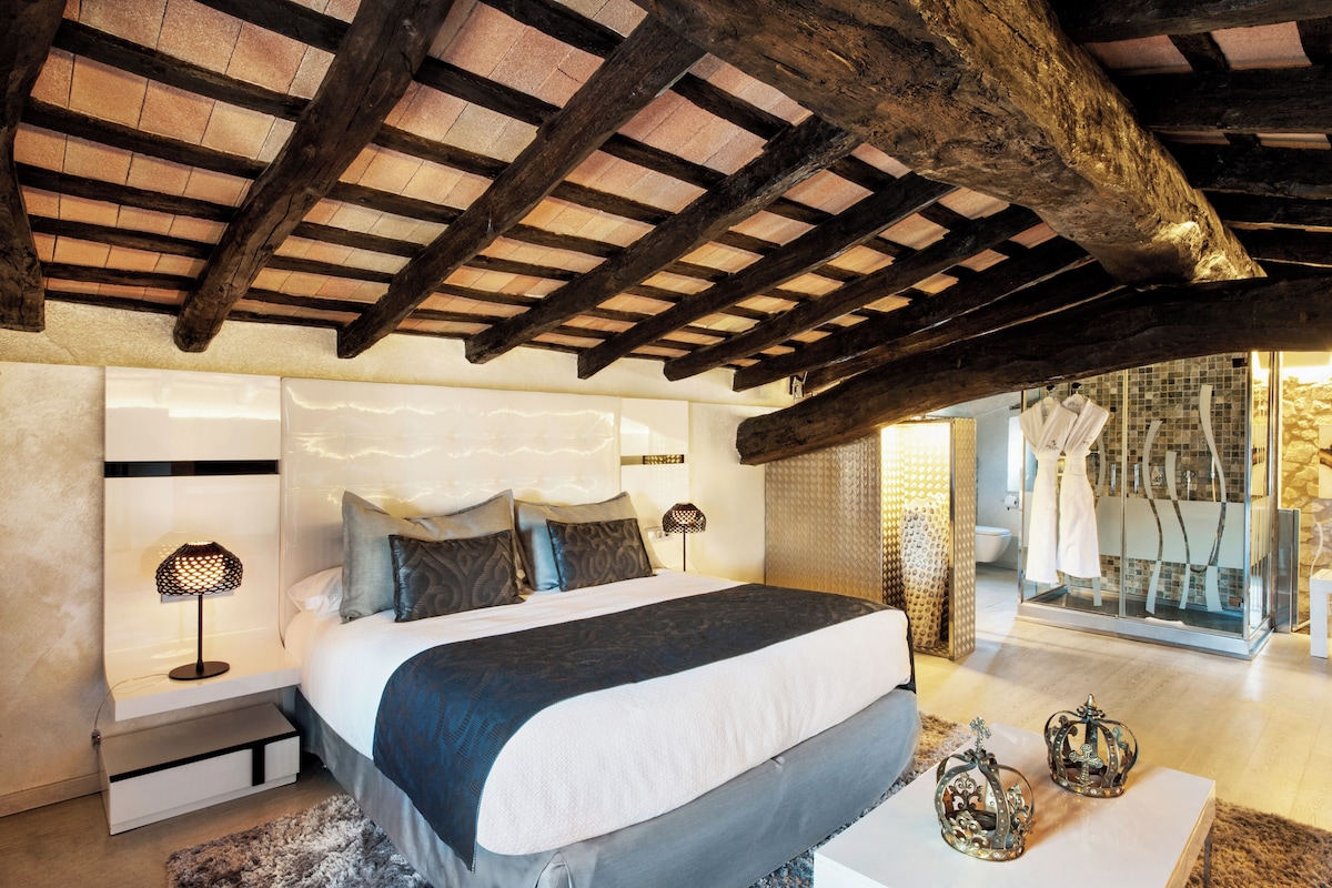 Un dormitorio con una cama grande, vigas de madera, cabecera moderna, lámparas de noche y un baño visible a través de una puerta de cristal.