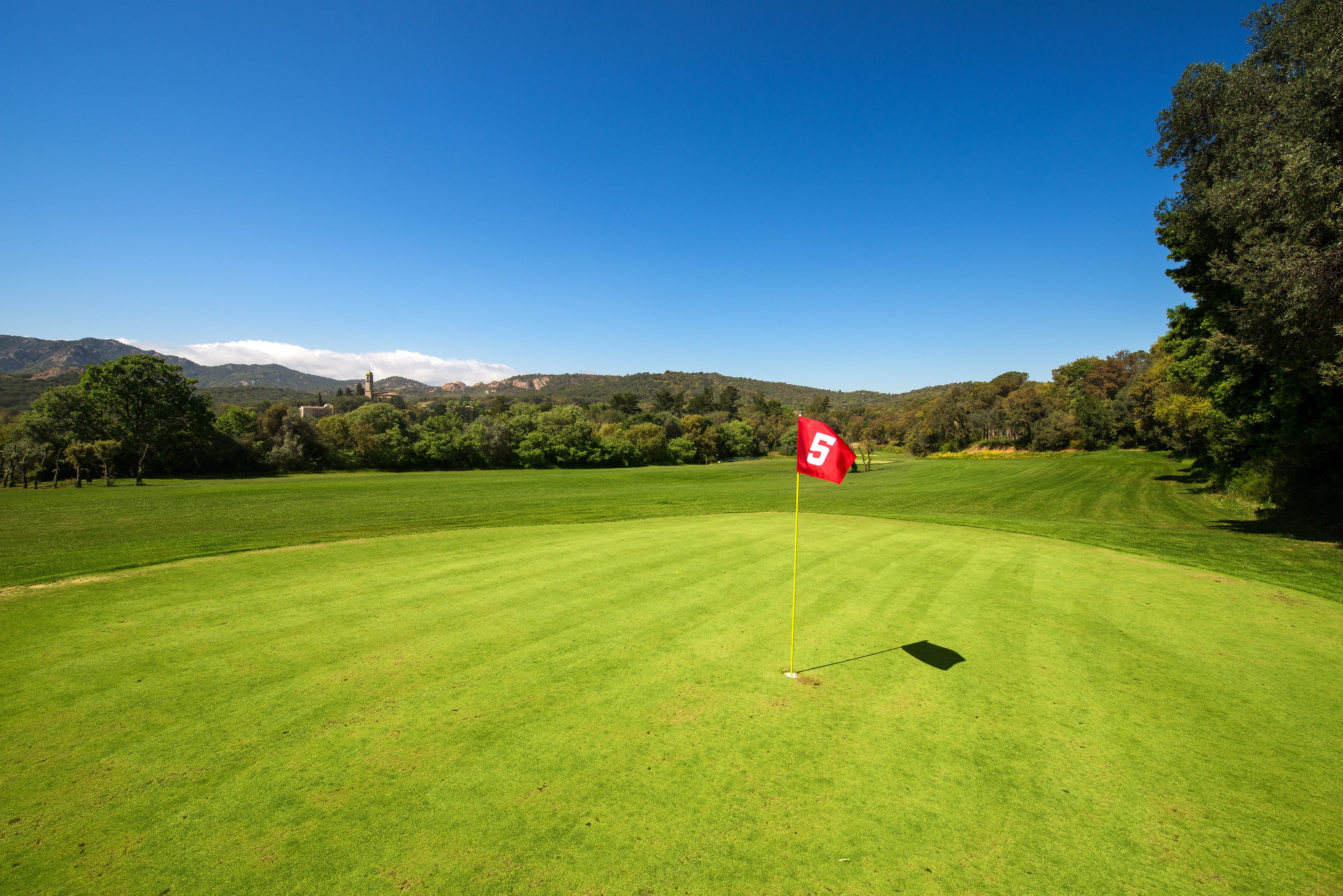Ein Golfplatz mit einer Fahne auf einem Abschlag, umgeben von Bäumen und Hügeln.