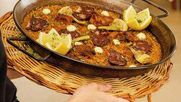 A person holding a large pan of paella with lemon slices and garlic cloves on a woven tray.