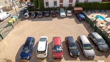 Un parking où de nombreuses voitures sont rangées en rangées, un petit espace piscine avec des chaises longues, et un bâtiment à la façade blanche, avec des espaces verts en arrière-plan.