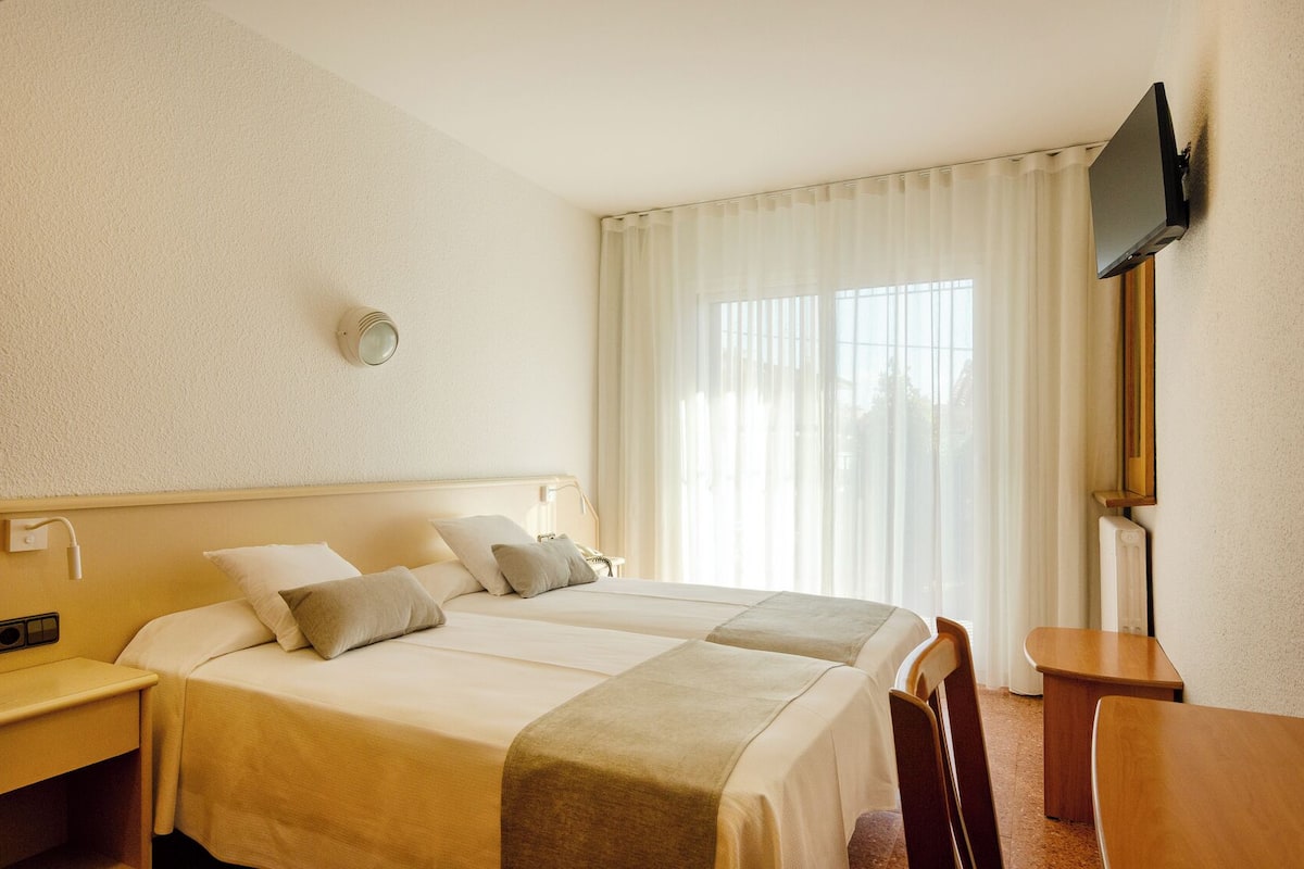 A hotel room with a bed, bedside tables, a desk, a chair, a flat-screen TV mounted on the wall, and a large window with curtains.