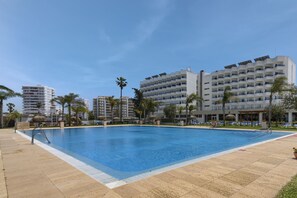 Piscina com água azul cristalina, rodeada por um deck revestido de azulejos. Há palmeiras e cadeiras de descanso ao redor da piscina.