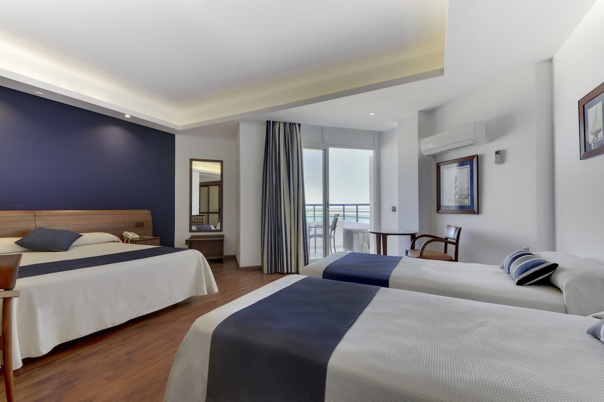 A hotel room with two beds, a desk, a chair, a window with curtains, and a view of the sea.