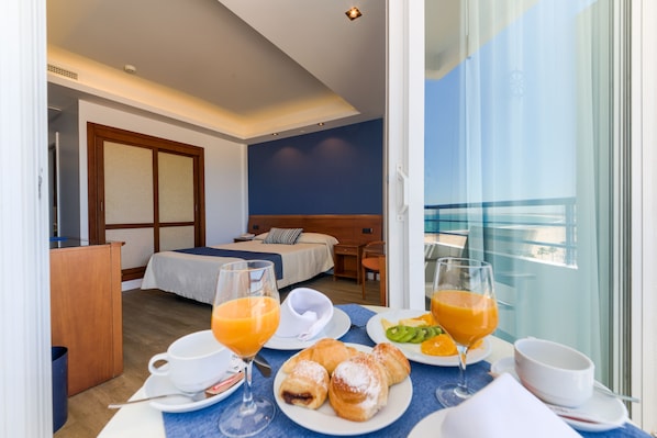 A hotel room with a bed, a desk, and a breakfast table with orange juice, croissants, and a bowl of fruit. - Hotel Puertobahia & SPA (El Puerto de Santa Maria)
