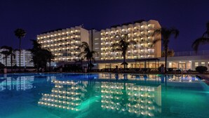 Um hotel com uma grande piscina que reflete as luzes do edifício, cercado por palmeiras.