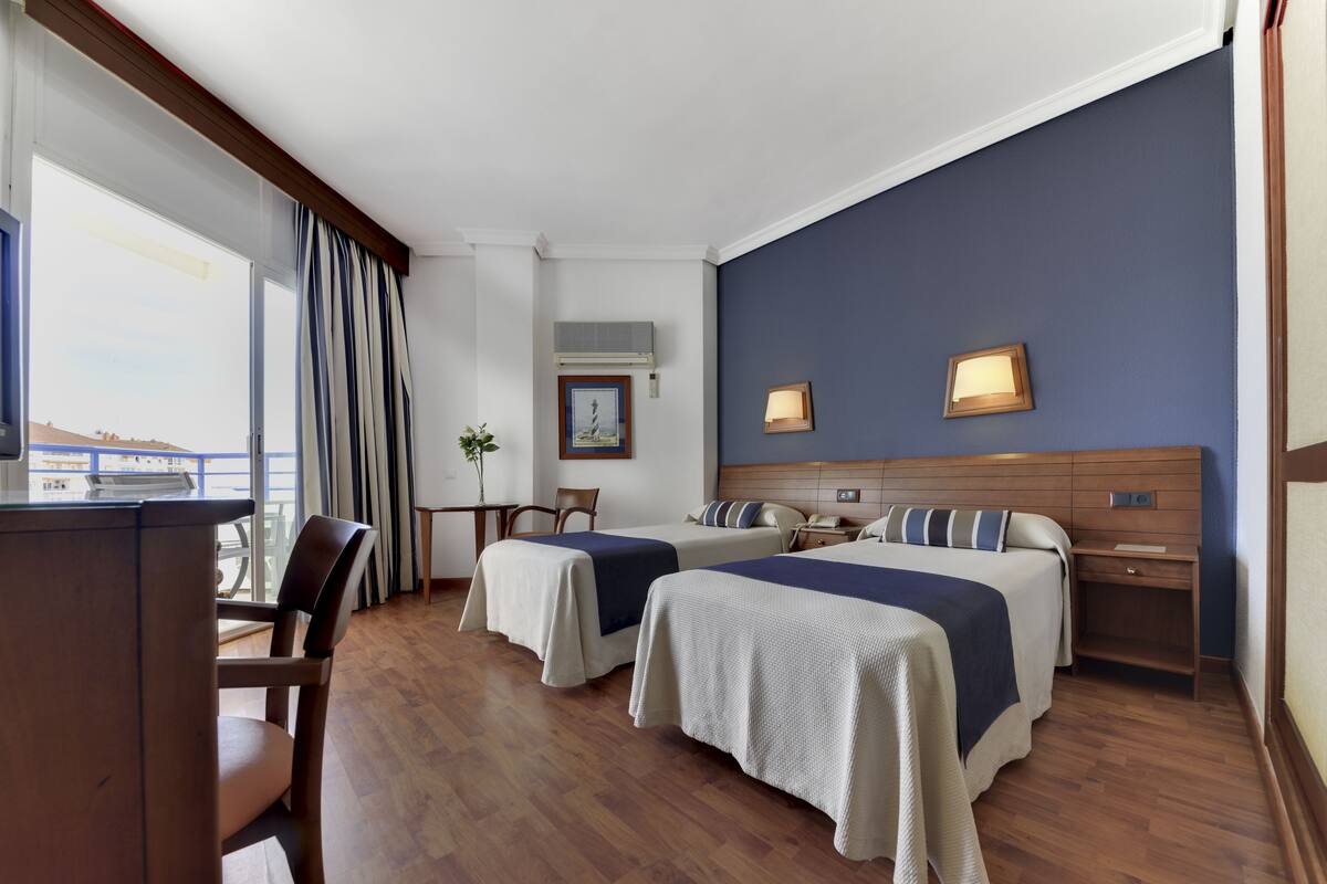 A hotel room with two beds, a desk, a chair, a window with curtains, and a view of the outside.