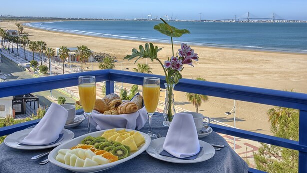 Mesa de café da manhã com pratos de frutas, croissants e suco de laranja, com vista para uma praia e uma ponte.