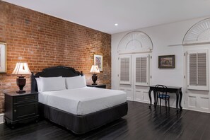 A bedroom with a brick wall, a large bed, two bedside tables with lamps, a chair, and a desk. - Hotel Royal (New Orleans)