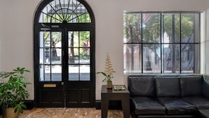 A room with a black door, a window with a view of the street, a black leather sofa, a small table with a plant, and a tiled floor. - Hotel Royal (New Orleans)