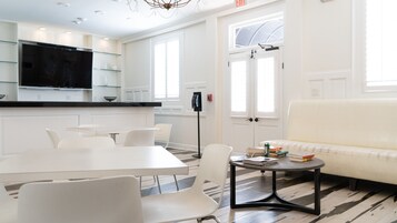 A modern living room with a white sofa, a dining table with chairs, and a kitchen area with a black refrigerator and glass shelves.