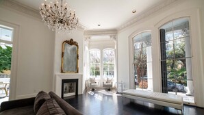A spacious living room with a dark wood floor, a large chandelier, and a bench by a large window. - Melrose Mansion (New Orleans)