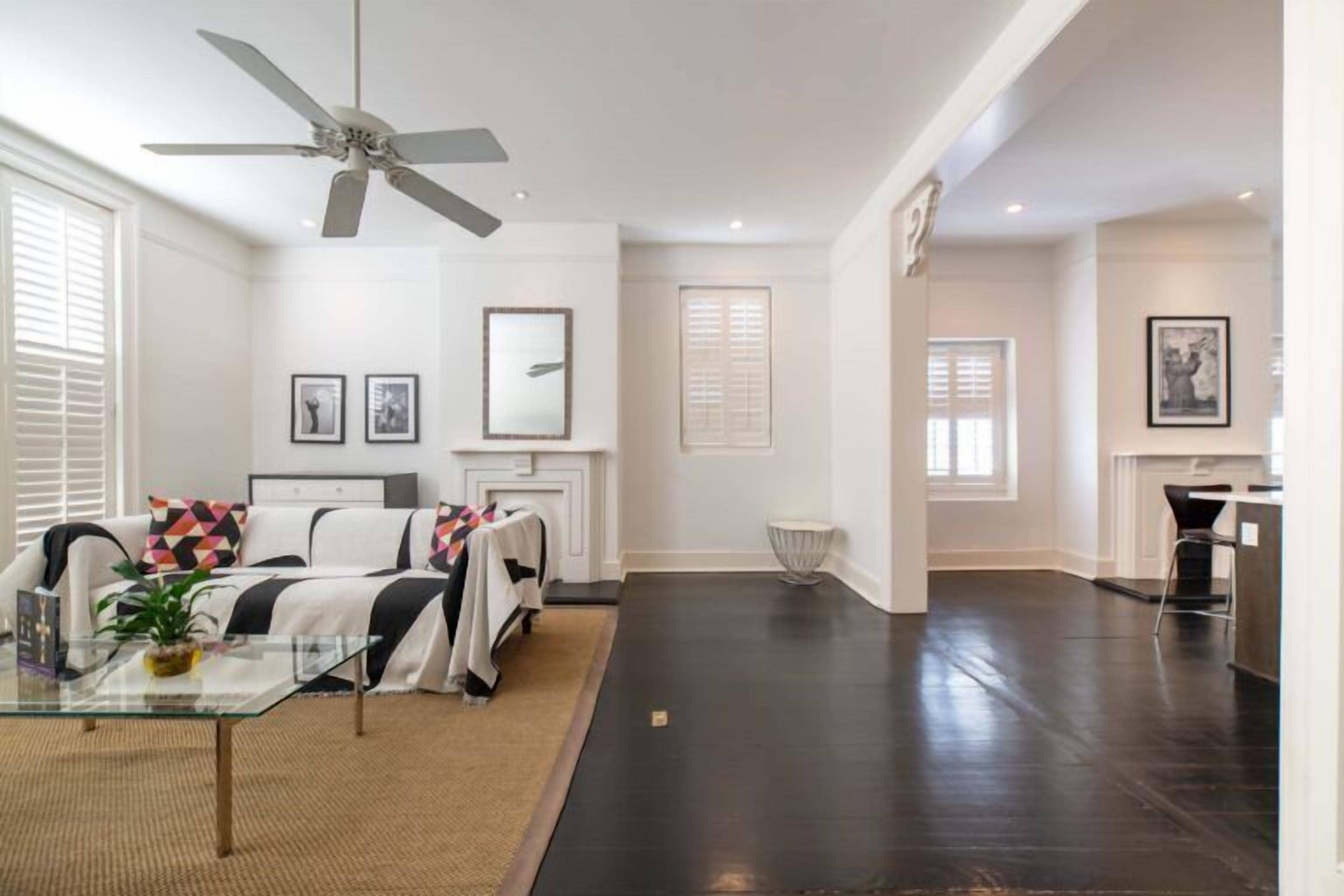 A spacious living room with a dark hardwood floor, a glass coffee table, a white sofa with colorful cushions, and a ceiling fan.