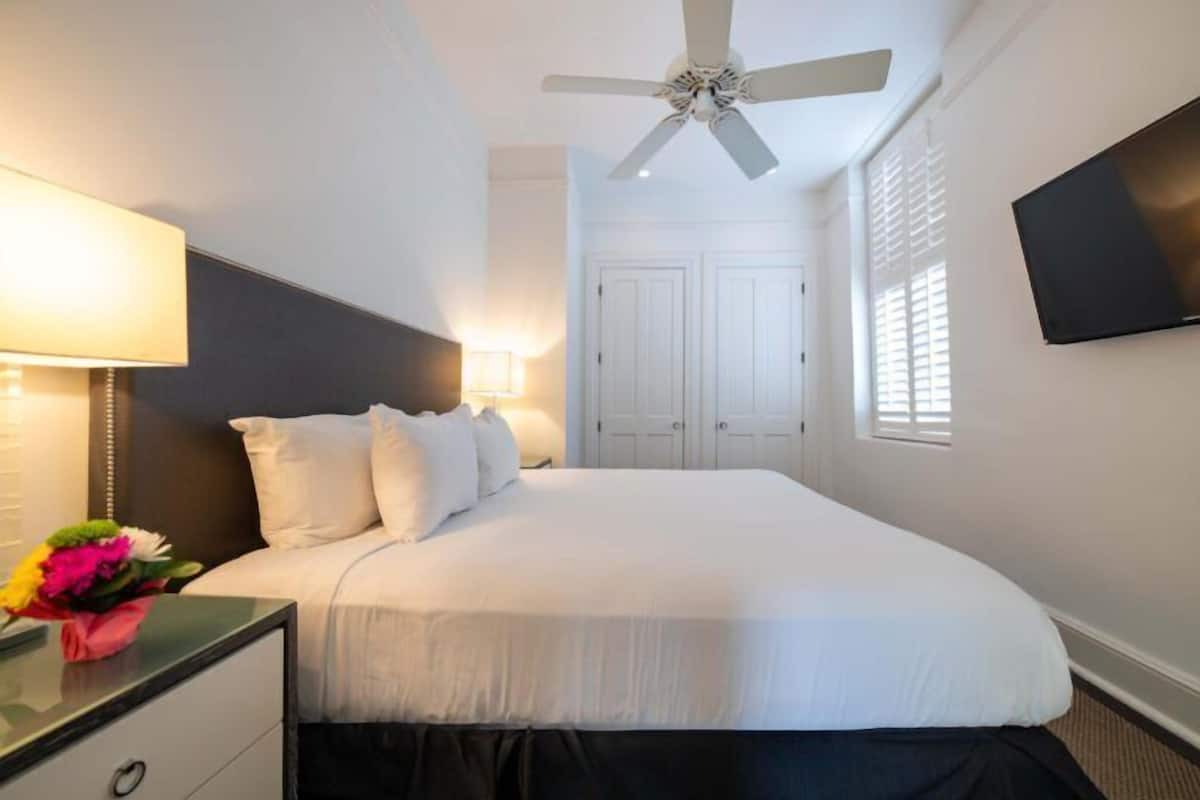 Une chambre d’hôtel avec un lit, un ventilateur de plafond, une télévision et une table de chevet avec une lampe et des fleurs.