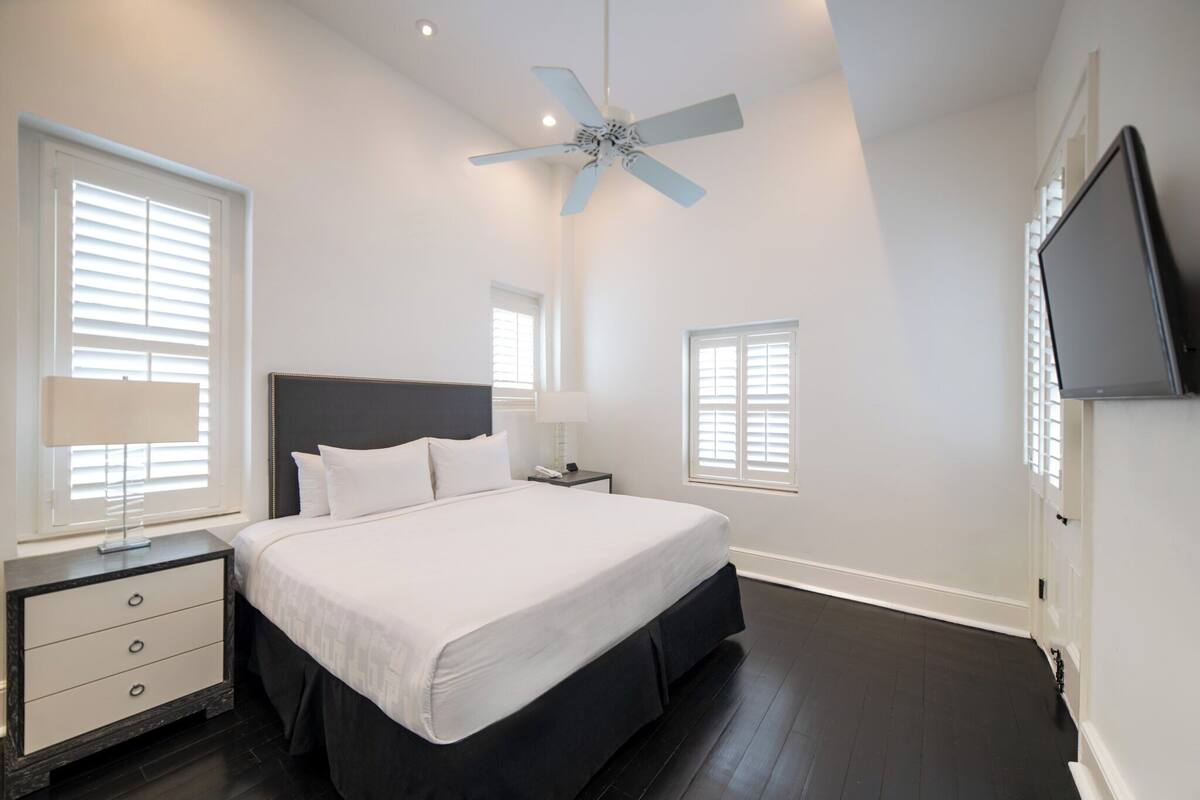 Une chambre à coucher avec un grand lit, une table de chevet, un bureau, un ventilateur de plafond et un téléviseur fixé au mur.