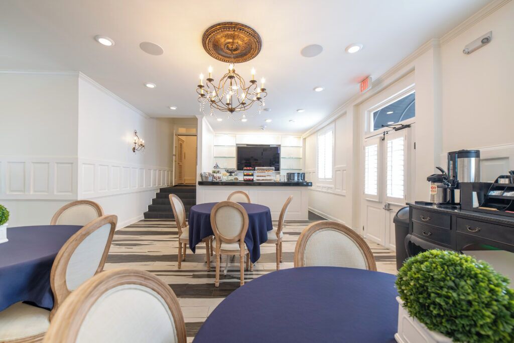 A hotel dining area with round tables, chairs, a chandelier, and a kitchen in the background.