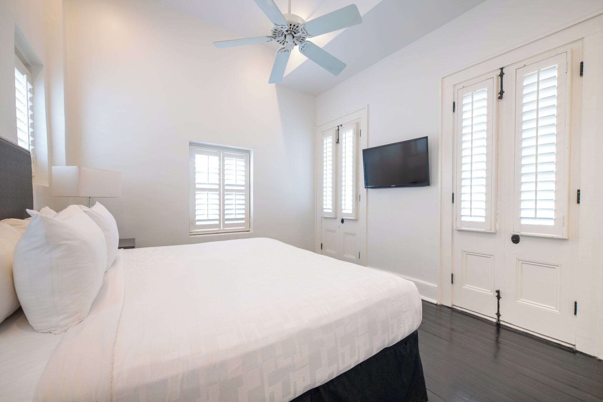 Un dormitorio con una cama grande, un ventilador de techo, un televisor en la pared y puertas blancas con ventanas.