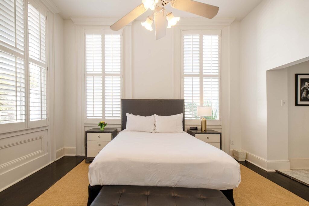 A bedroom with a large bed, two bedside tables, a ceiling fan, and large windows.