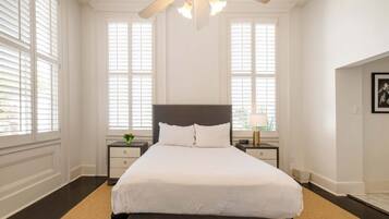 Un dormitorio con una cama grande, dos mesitas de noche, un ventilador de techo y ventanas amplias.