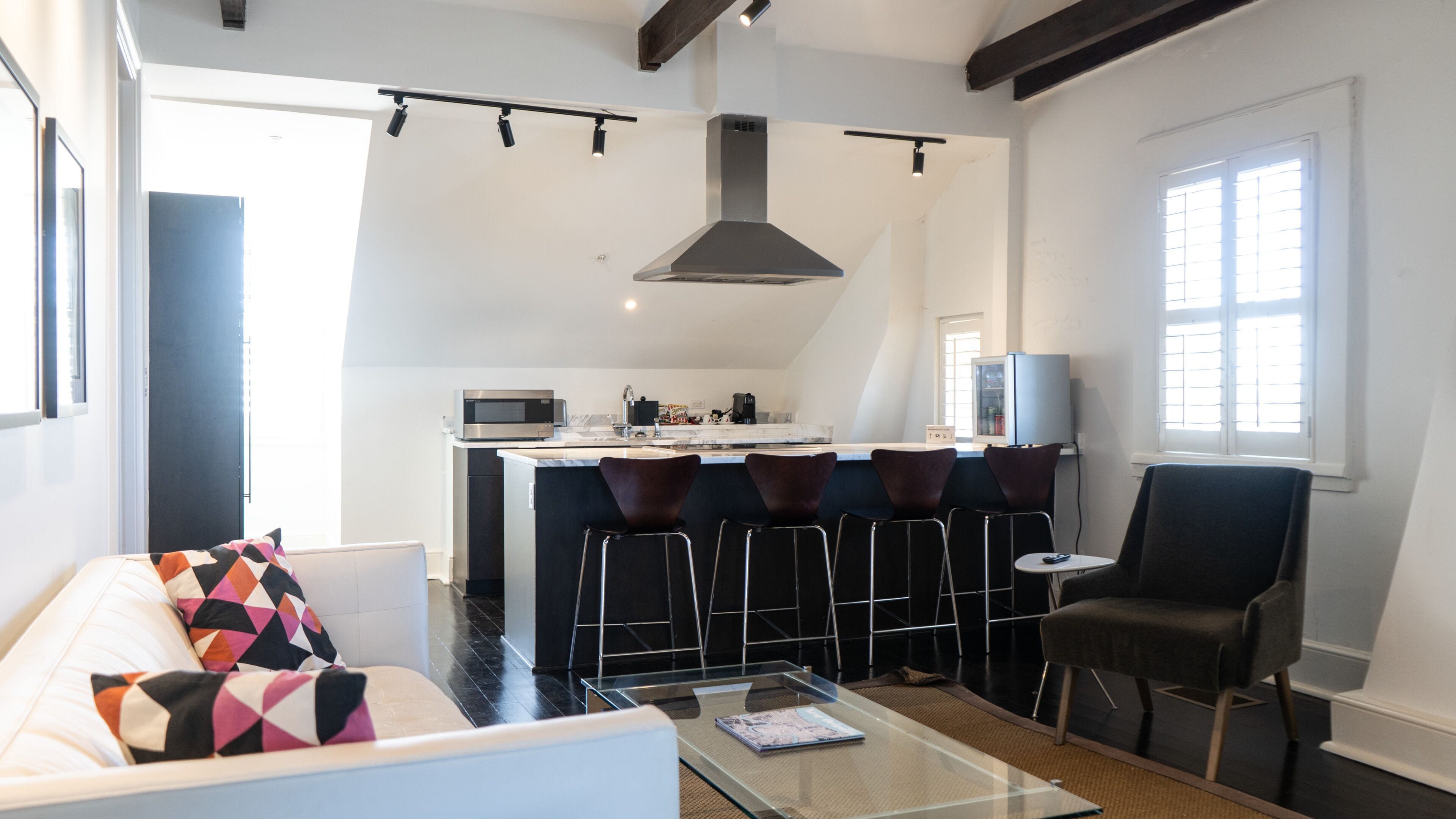 A modern living room with a glass coffee table, a sofa with patterned cushions, a black armchair, and a kitchen area with a microwave, stove, and bar stools.