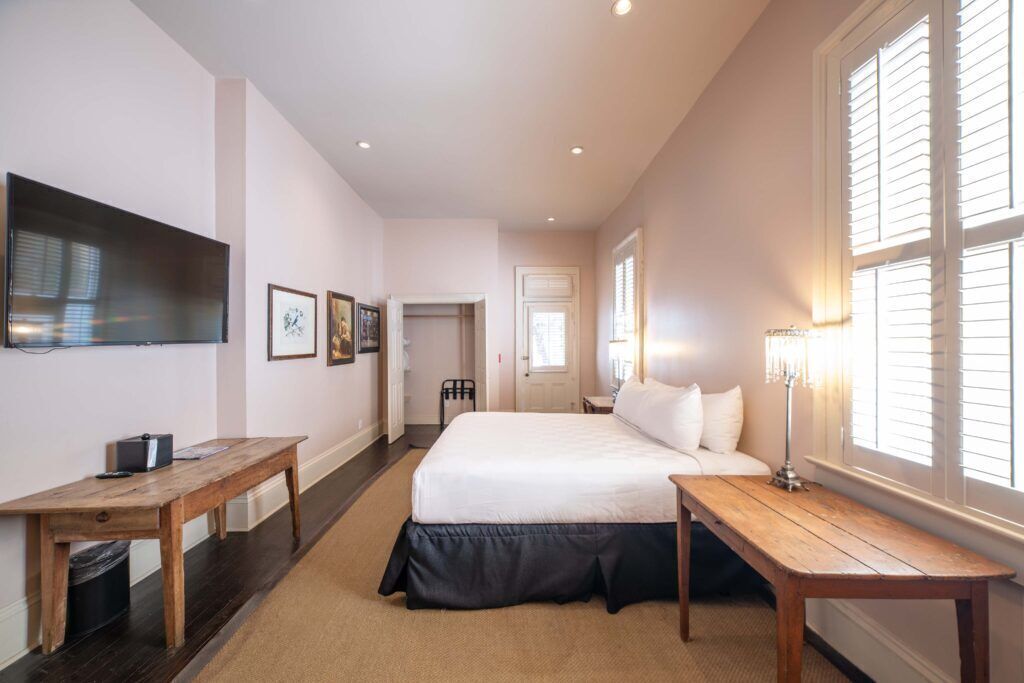 A bedroom with a large bed, a wooden desk, a TV mounted on the wall, and a window with blinds.
