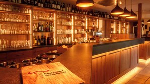 Un bar restaurante con estantes de madera llenos de botellas y vasos, una barra con una carta y luces colgantes.