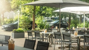 Un café al aire libre con mesas y sillas bajo sombrillas, rodeado de vegetación y coches aparcados.