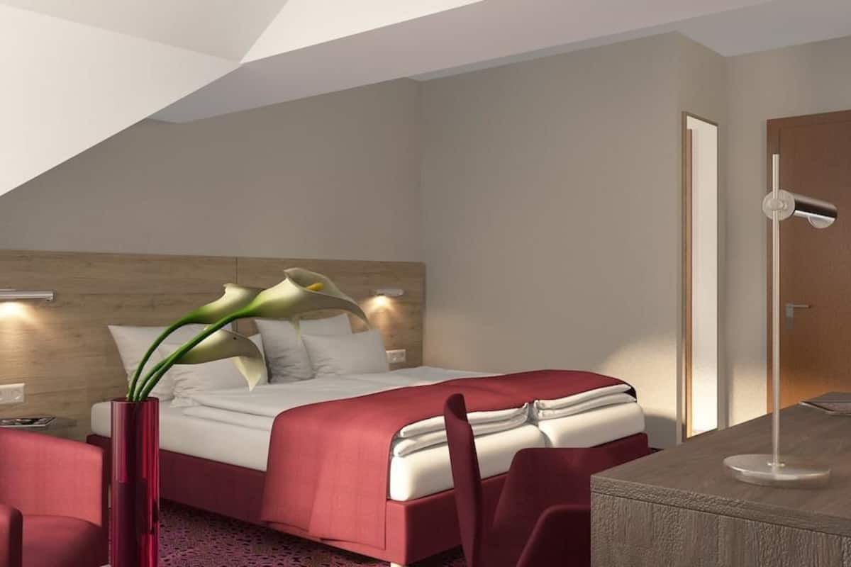A modern hotel room with a bed, a desk, a chair, a vase with flowers, and a tall floor lamp.