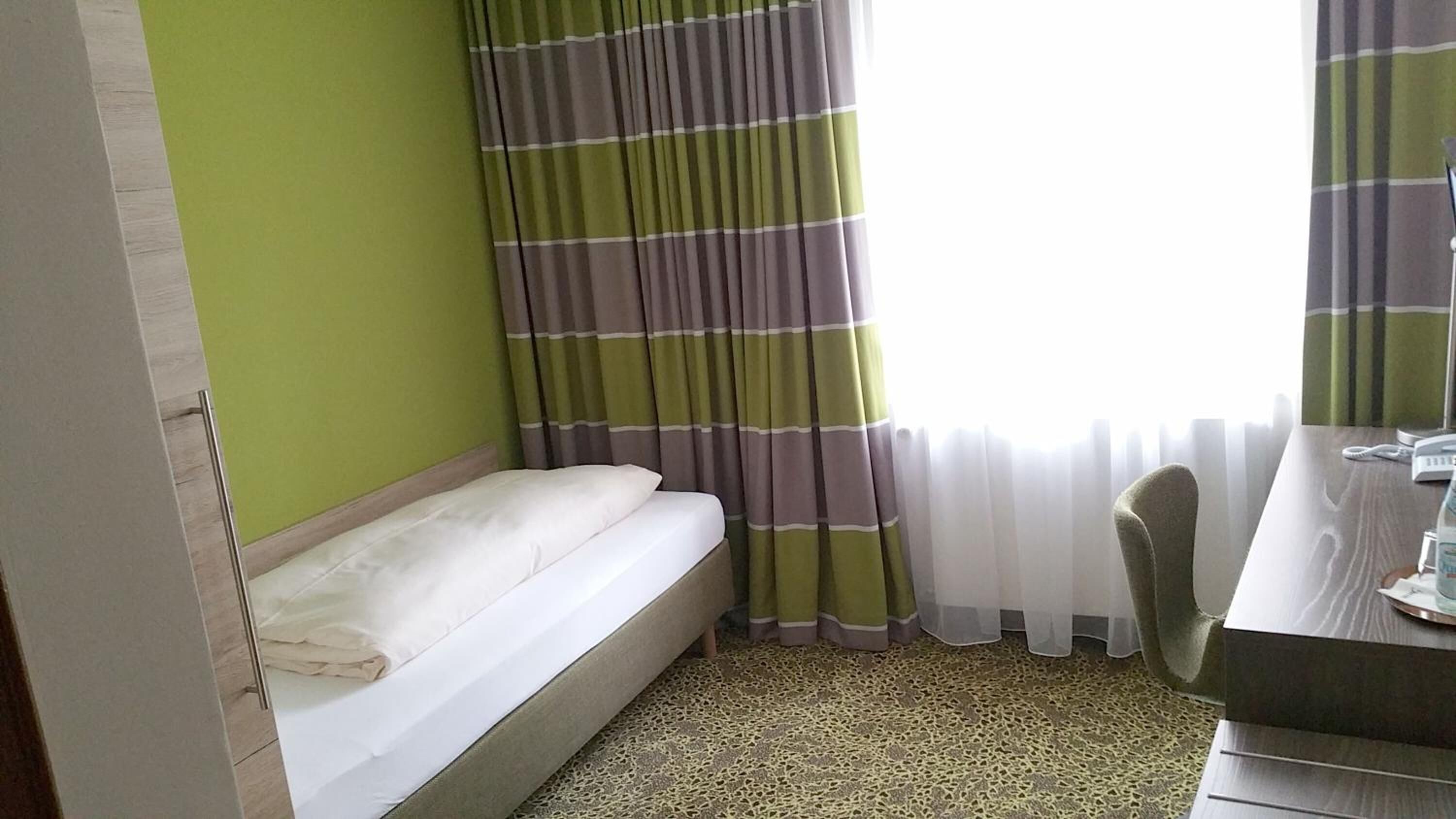 A small, single-bed room with a green wall, a window with striped curtains, a desk with a chair, and a telephone.