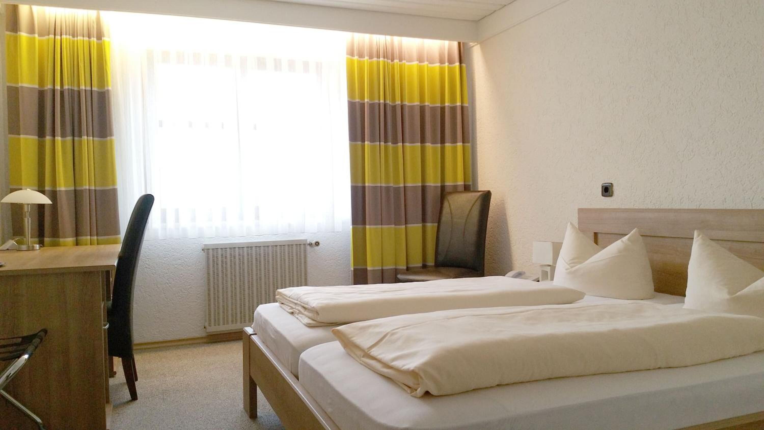 A hotel room with two beds, a desk with a lamp, a chair, and a radiator.