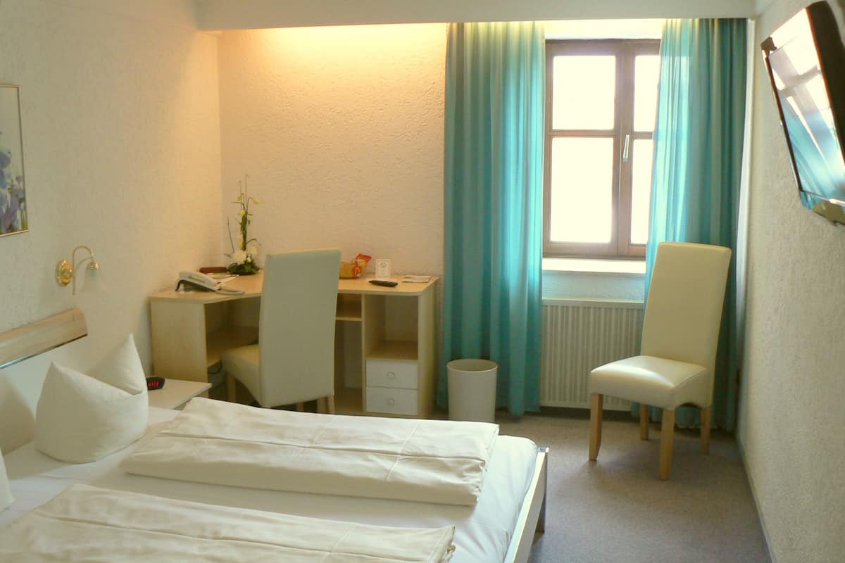 A hotel room with a bed, a desk, a chair, a radiator, and a window with curtains.